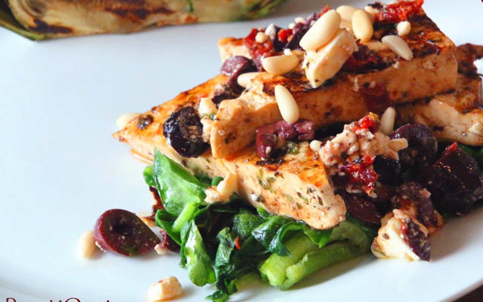 Mediterranean Tofu Steaks [Vegan, Gluten-Free] over bed of spinach with dairy-free feta and pine nut topping